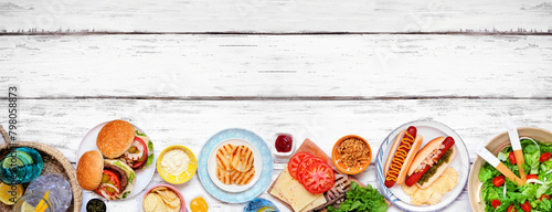 Summer BBQ or picnic bottom border with hamburgers, hotdogs, salad and snacks. Top down view over a white wood banner background. Copy space.