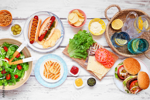 Summer BBQ or picnic table scene with hamburgers, hotdogs, salad and snacks. Overhead view on a white wood background.