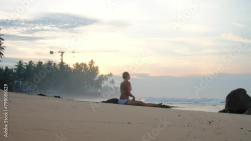 Woman in bikini engages in yoga session on sandy beach near ocean. Lady practices sitting straddle asana and mist covers warm water