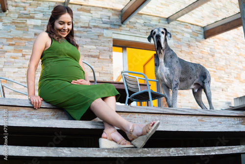 pregnant woman relaxes sitting on the porch of her house next to her dog. she happily looks at and caresses her tummy belly.
