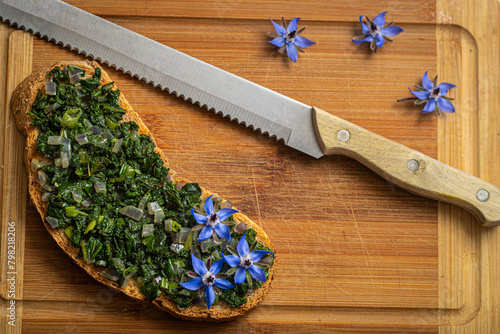 A crispy bruschetta, generously topped with borage and onion, served on rustic bread. A wooden-handled knife suggests freshness.