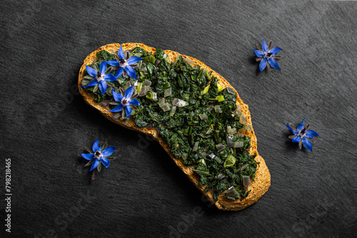 Vibrant blue flowers and fresh borage elegantly decorate a crispy bread slice, showcased against a striking black background