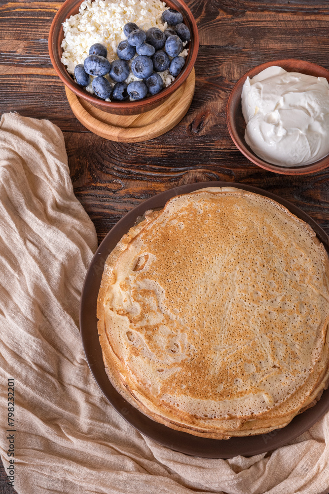 A plate of golden pancakes topped with blueberries, accompanied by a bowl of sour cream, a portion of fresh cottage cheese and a glass of milk. On a beige-golden background with a green runner. A plate of golden pancakes topped with blueberries, accompanied by a bowl of sour cream, a portion of fresh cottage cheese and a glass of milk. On a beige-golden background with a green runner.