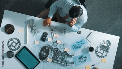Top view of engineer talking to phone while working on tablet with green screen at meeting table with turbine engine or jet engine sketch design and various metal gear scatter around. Alimentation.