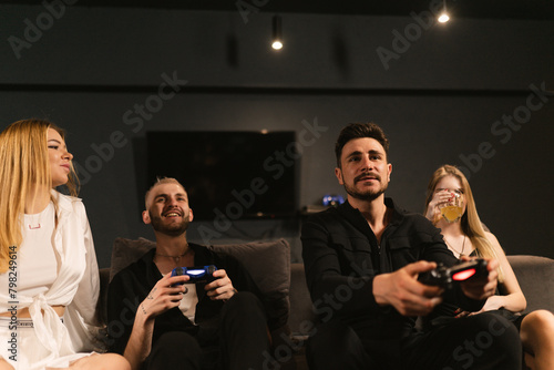 Young men delve into virtual combat on console sitting on sofa with girlfriends. Women actively support guys savoring drinks