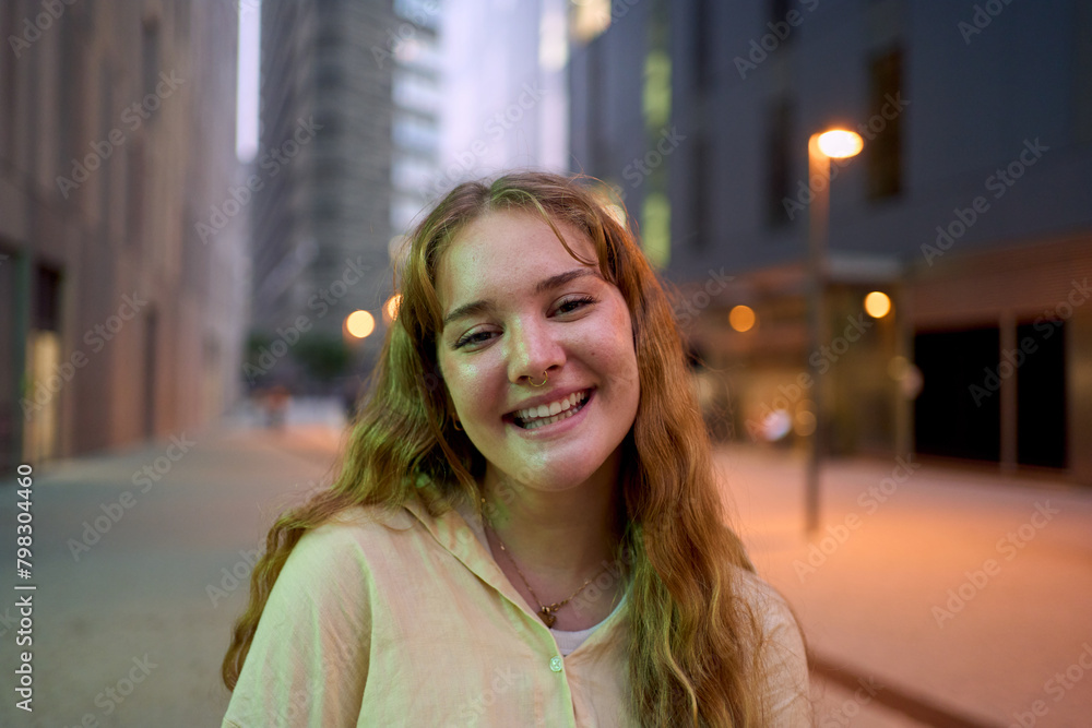 Portrait nice Caucasian young girl smiling looking happy at camera urban night outdoors. Generation z friendly only blonde woman posing cheerful in street with city light in background. Copy space 
