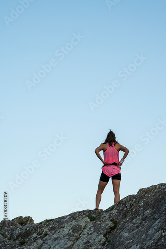 A girl stands on top of a mountain and prepares to run down the mountain. She is dressed in a black shirt and a pink shirt. 