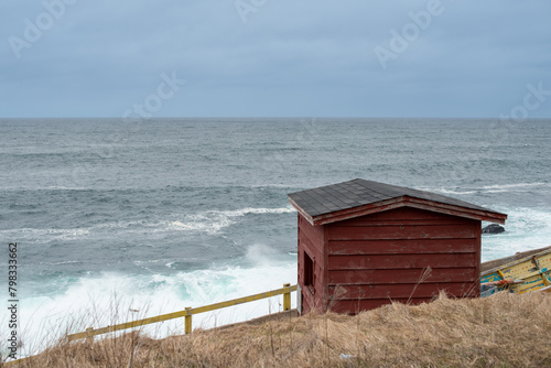 A small red wooden worn and weathered fishing shed or boat house. The exterior has red boards and a small window with a black roof. There's a stormy sea in the background with large waves and blue sky