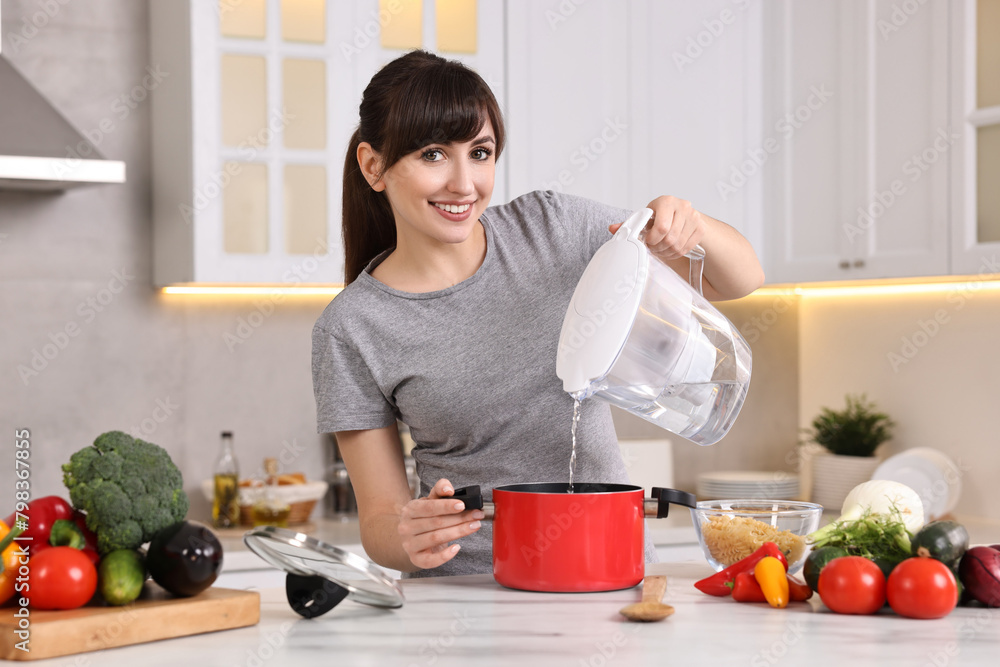 Happy young housewife pouring water in pot at white marble table in kitchen. Cooking process