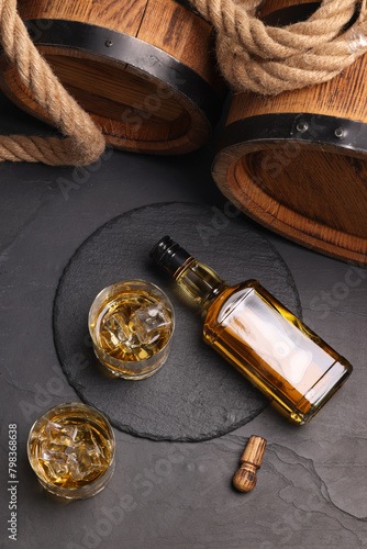 Whiskey with ice cubes in glasses, bottle, wooden barrels and rope on black table, flat lay