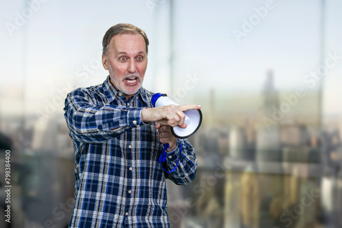 Confident elderly mature man giving a speech in megaphone and pointing at something. Boss giving a command. Blurred indoors background. photo