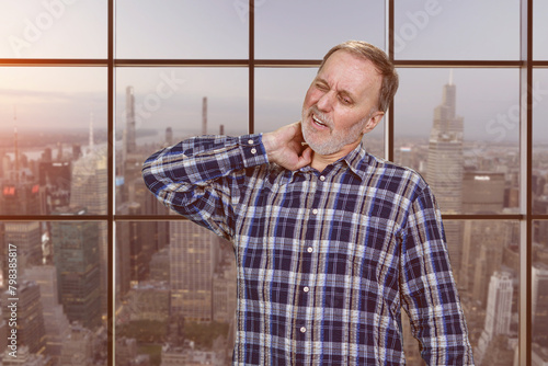 Handsome mature caucasian man is having a neckache. Checkered windows background with cityscape view.