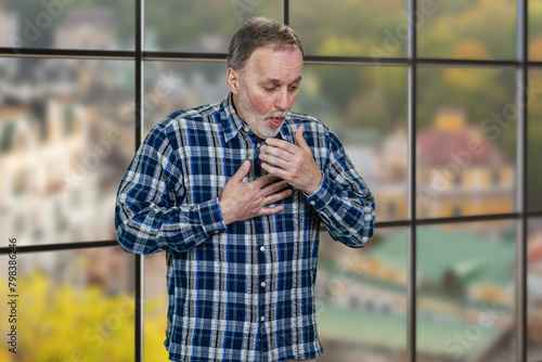 Portrait of a mature man coughing standing indoors. Windows background with cityscape view. © DenisProduction.com