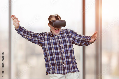 Portrait of excited old man in headset with his arms spreaded. Blurred windows background. © DenisProduction.com