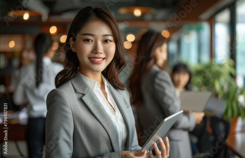 Smiling confident business leader looking at camera holding a digital tablet and standing in office at team meeting. Portrait of pretty Asian businesswoman with colleagues. AI Generated © EarthWalker