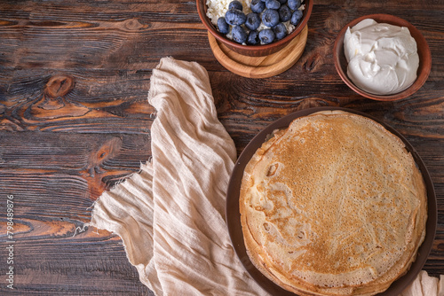A plate of golden pancakes topped with blueberries, accompanied by a bowl of sour cream, a portion of fresh cottage cheese and a glass of milk. On a beige-golden background with a green runner.