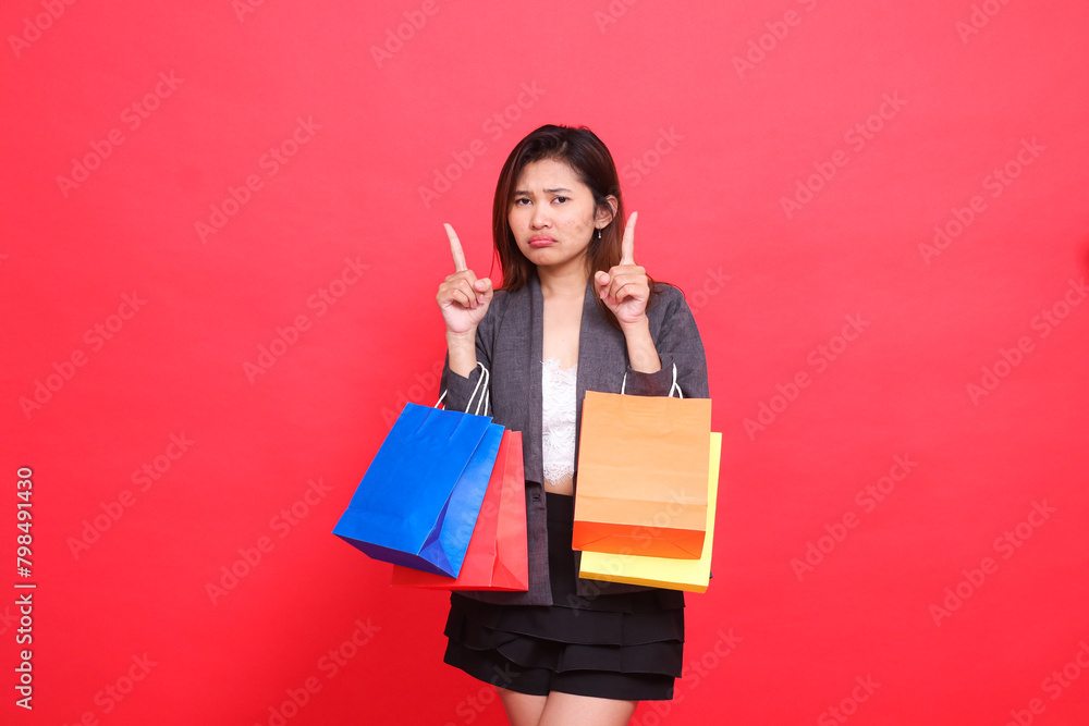 the expression of a sad Asian office woman carrying shopping paper bags with both hands pointing upwards wearing a formal jacket on a red background. for commercial, lifestyle and sales concepts the expression of a sad Asian office woman carrying shopping paper bags with both hands pointing upwards wearing a formal jacket on a red background. for commercial, lifestyle and sales concepts
