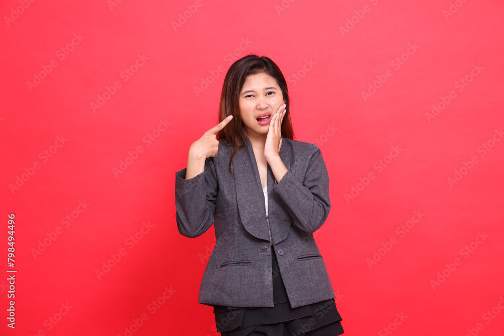 the expression of an Asian office woman in pain with a toothache while pointing at her wearing a gray jacket and black mini skirt on a red background. for health, business and advertising concepts the expression of an Asian office woman in pain with a toothache while pointing at her wearing a gray jacket and black mini skirt on a red background. for health, business and advertising concepts