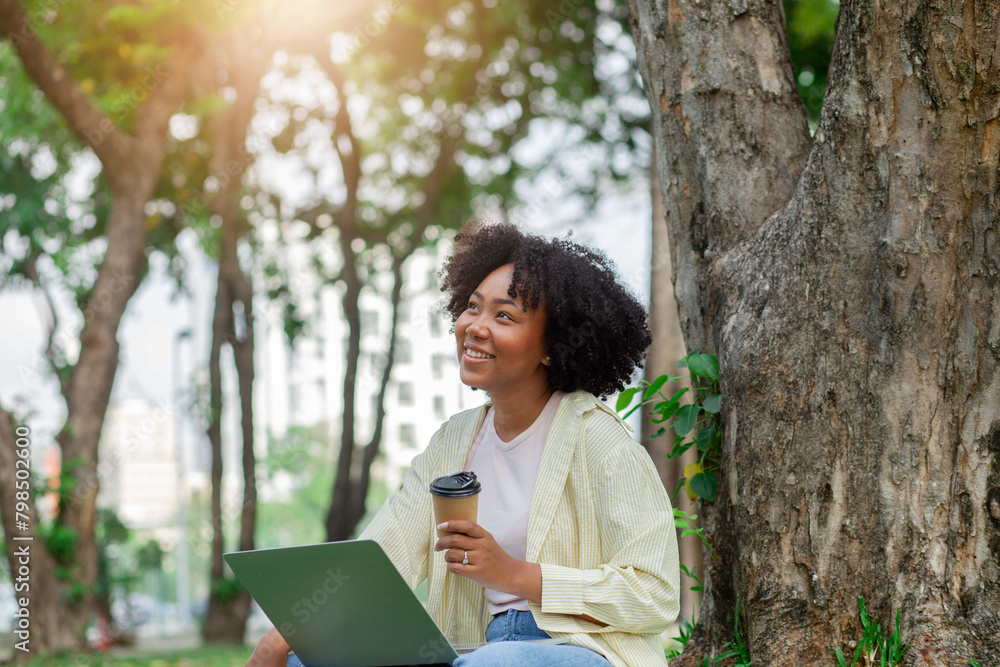 A woman is sitting on a tree stump with a laptop and a cup of coffee. She is smiling and seems to be enjoying her time in the park