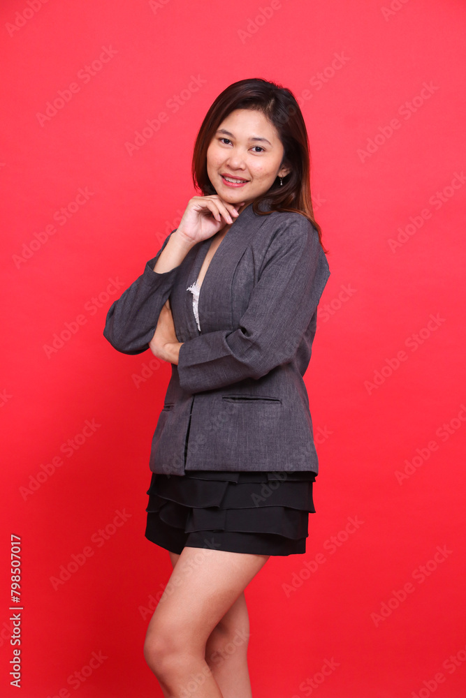 Portrait of a cheerful Asian office woman with her right arm crossed, holding her chin, wearing a gray jacket and black mini skirt on a red background. for corporate concepts, branding and advertising Portrait of a cheerful Asian office woman with her right arm crossed, holding her chin, wearing a gray jacket and black mini skirt on a red background. for corporate concepts, branding and advertising