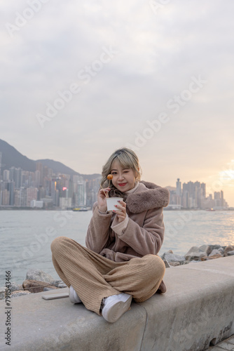 A young woman in winter clothes in her 20s who eats local food fish balls in the evening in a park with a view of buildings and the sea on Kowloon Island, Hong Kong
香港九龍島のビル群と海が見える公園で夕方にローカルフードのフィッシュボ