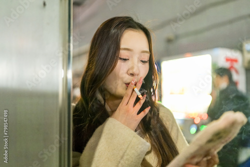 A Chinese woman in her 20s smoking while operating her smartphone at a smoking area in front of the station in Omiya Ward, Saitama City, Saitama Prefecture 埼玉県さいたま市大宮区の駅前の喫煙所でスマートフォンを操作しながらタバコを吸う２０代の中