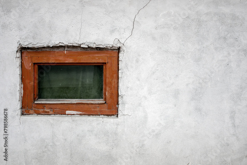 Lonely window. Destroyed plaster on an old white wall of a house. A small window with a brown frame on a white plastered wall.