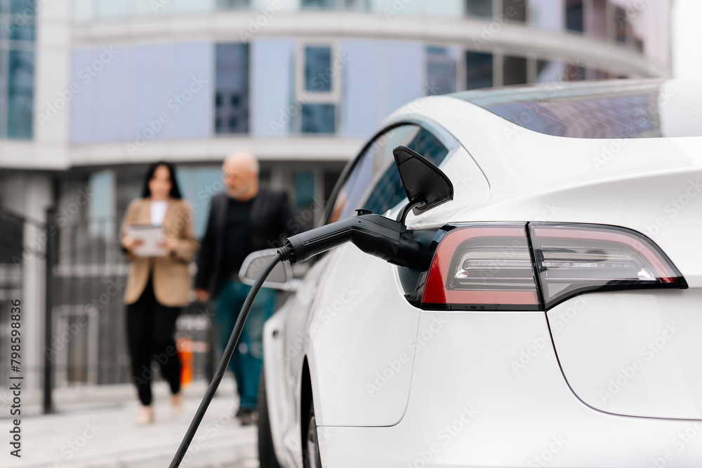 An unrecognizable modern electric car is charging. In the background, two business people, a man and a woman, sign a contract near a business center. An unrecognizable modern electric car is charging. In the background, two business people, a man and a woman, sign a contract near a business center.