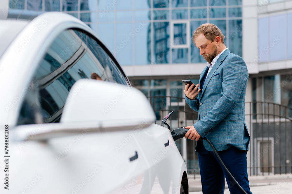 A middle-aged businessman charges an electric car from a charging station powered by renewable energy sources against the backdrop of the business center. A middle-aged businessman charges an electric car from a charging station powered by renewable energy sources against the backdrop of the business center.