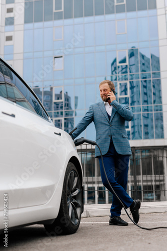 A young successful businessman in a stylish suit uses a telephone against the backdrop of a modern business center and a modern electric machine. Concept of electric vehicles, business and style. © Vladislav