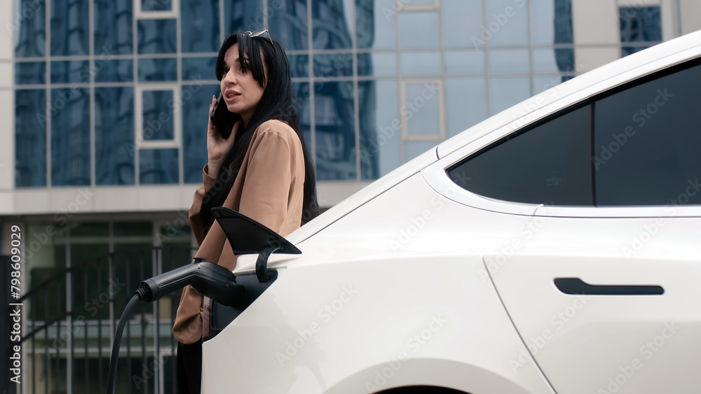 Close-up of a modern electric car charging against the backdrop of a business woman and a modern business center. Concept of using energy, electrical machines and modern technologies.