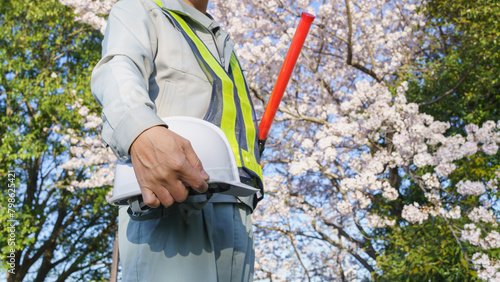 春の桜を背景に誘導棒とヘルメットを持つ作業服の男性｜建設業・警備業などのイメージ