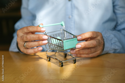 woman's hands hold a miniature supermarket cart. The woman is wearing a blue shirt. Groomed hands of an elderly woman. Concept of finance and shopping.