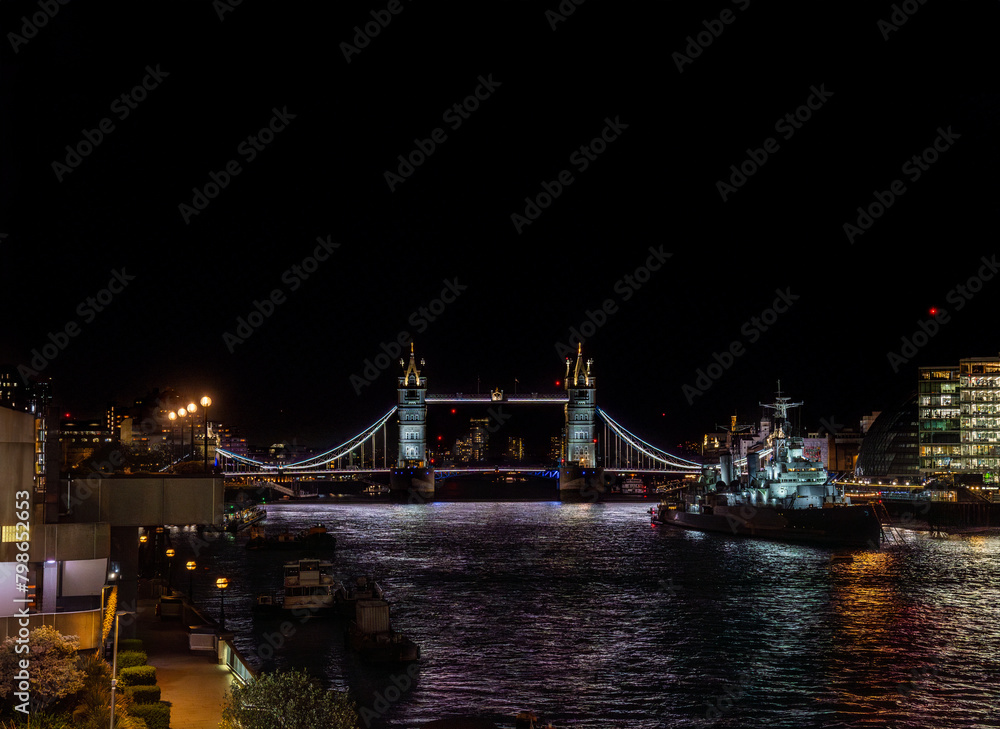 Front view of Tower Bridge illuminated at night and the war museum ship HMS Belfast docked on the River Thames with the lights of restaurants, buildings reflected in the water.