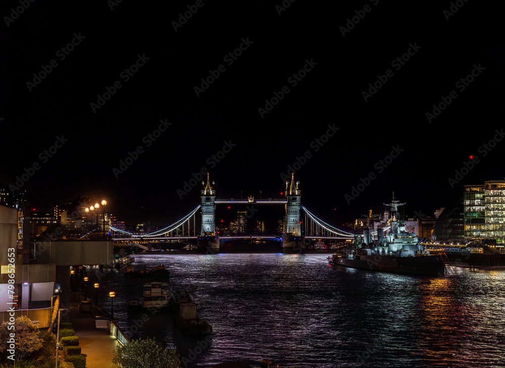 Obraz premium Front view of Tower Bridge illuminated at night and the war museum ship HMS Belfast docked on the River Thames with the lights of restaurants, buildings reflected in the water.