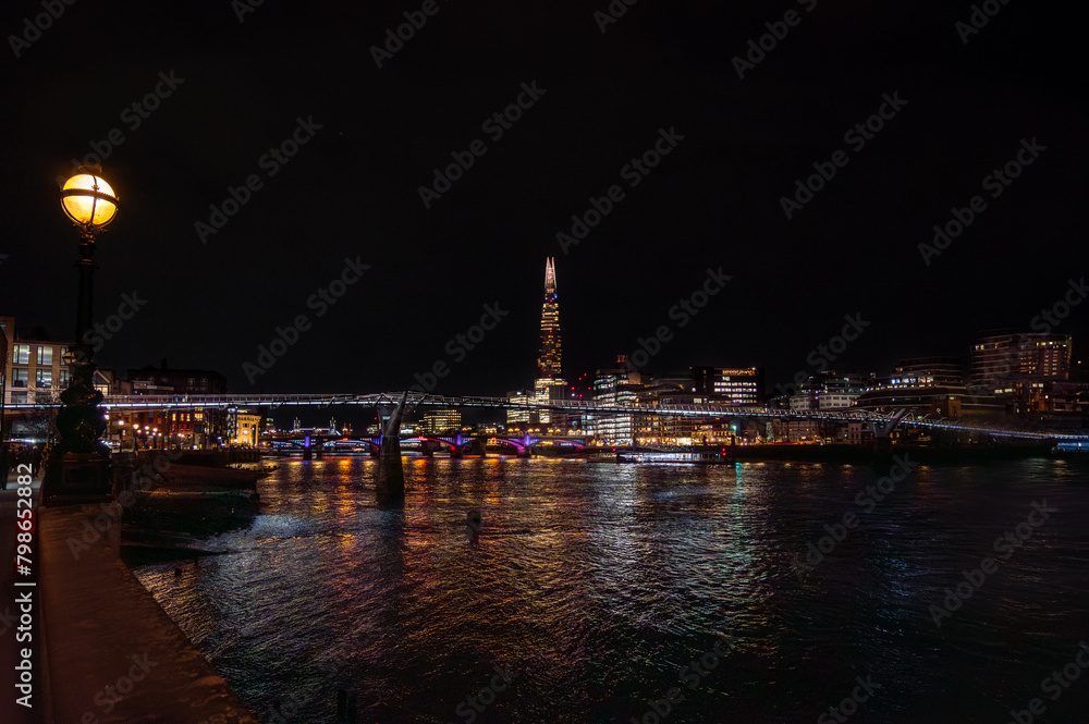 The River Thames illuminated by the reflections of colored lights from the buildings, promenade with street lamps and The Shard skyscraper and the Millennium Bridge and Tower Bridge in the background. The River Thames illuminated by the reflections of colored lights from the buildings, promenade with street lamps and The Shard skyscraper and the Millennium Bridge and Tower Bridge in the background.