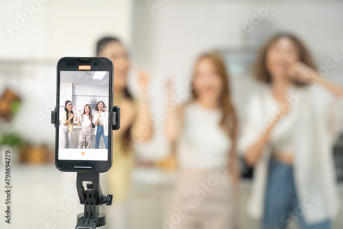 Smartphone on a tripod recording a lively moment of three friends laughing and enjoying themselves in a kitchen.