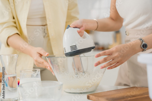 Hands of two people using an electric mixer to blend ingredients in a bowl during a home baking session.