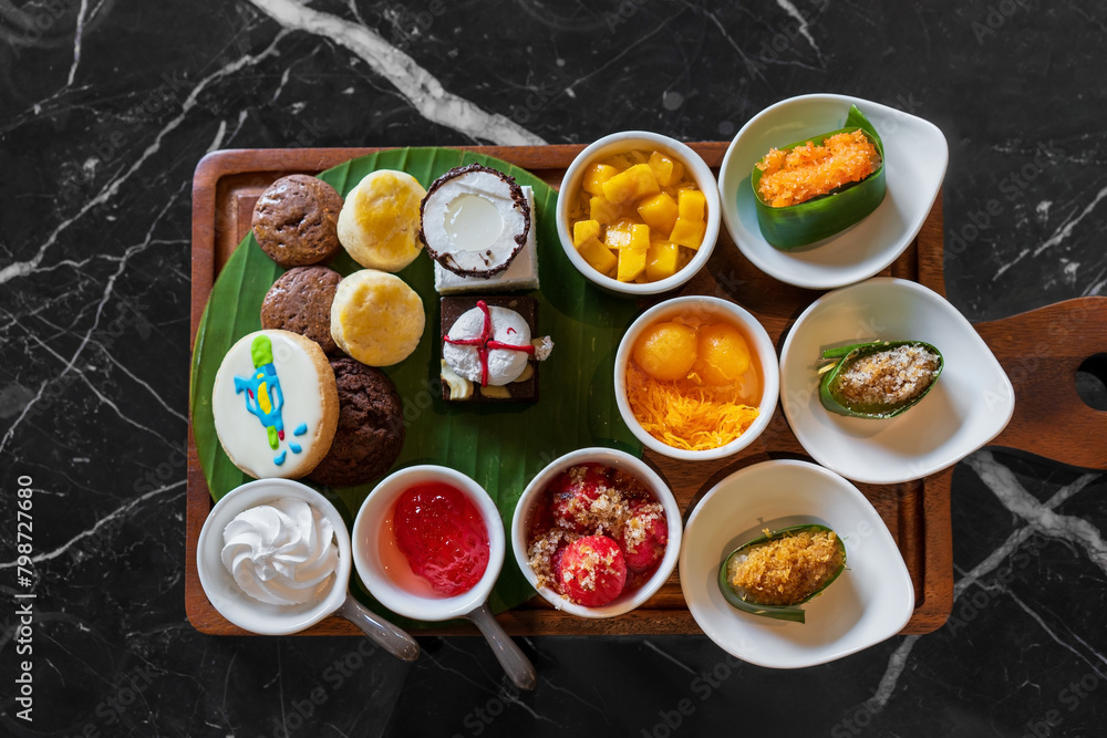 Set various types of Thai desserts in small bowls with thong yip thong yot cake cookies chocolate and mango sticky rice on banana leaf and top view black marble table for sweet snacks food at bakery