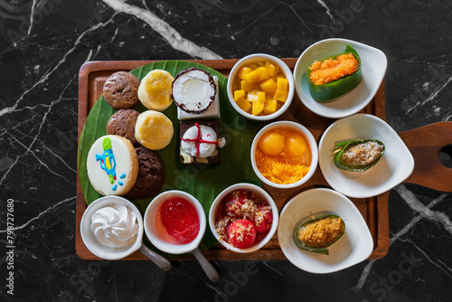Set various types of Thai desserts in small bowls with thong yip thong yot cake cookies chocolate and mango sticky rice on banana leaf and top view black marble table for sweet snacks food at bakery