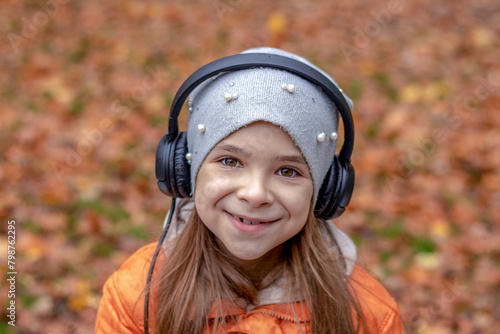 A girl listens to music on headphones in an autumn park and looks at the camera.