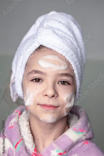 A girl with a white bath towel on her head and cleansing foam applied to her face smiles and looks at the camera.