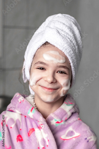 A girl with a towel on her head in the bathroom washes her face with cleansing foam.