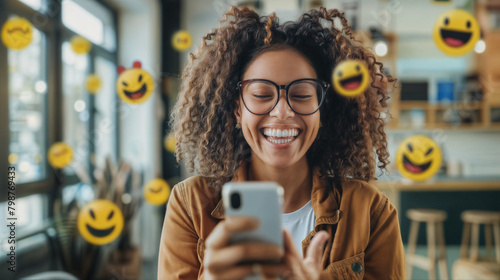 A young smiling black woman holding a mobile phone in the office with emoji smiles flying around her. The person receives a pleasant message photo