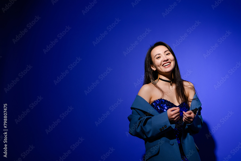 Cheerful and stylish asian woman in dress and jacket posing on blue background Cheerful and stylish asian woman in dress and jacket posing on blue background
