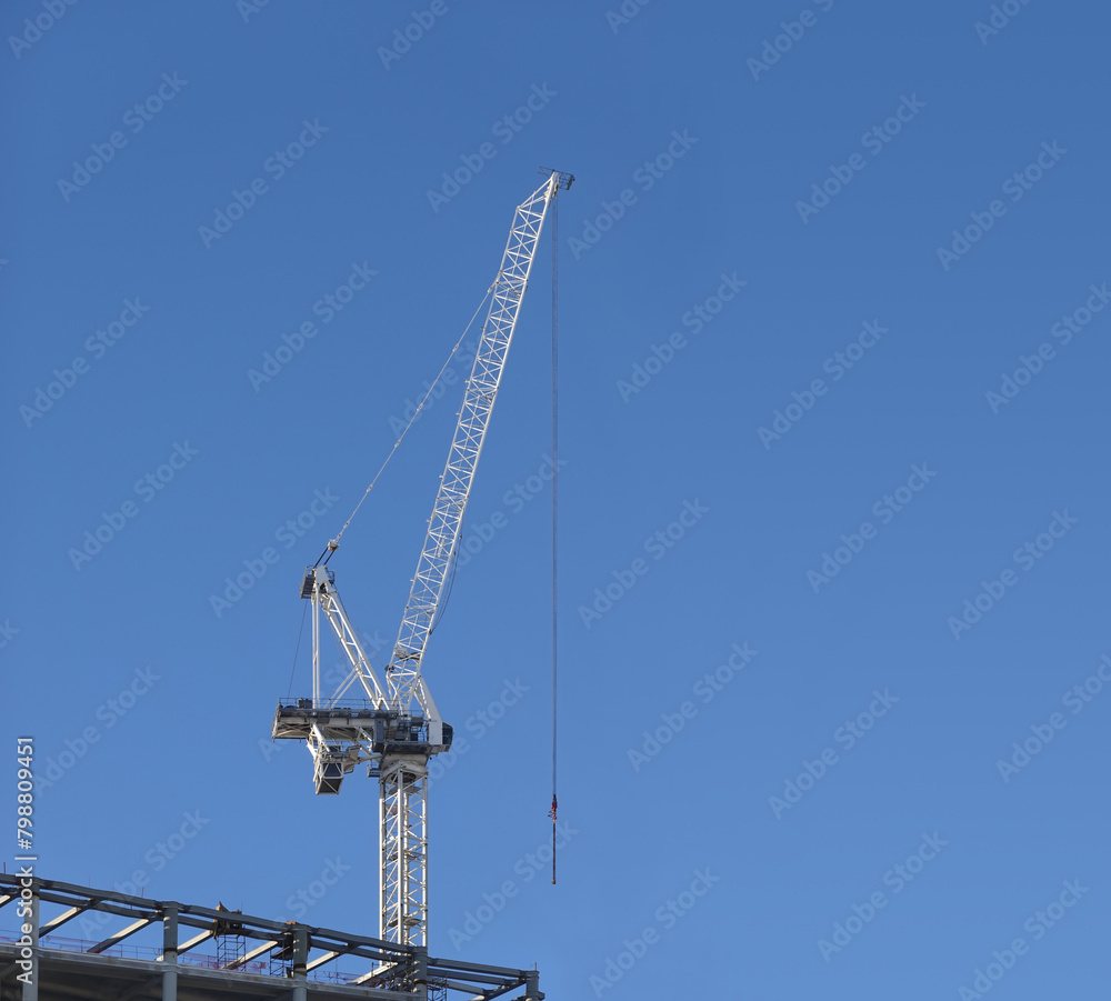 Tall hoisting tower crane on the top section of being constructed of modern high skyscraper building against clear blue cloudless sky Tall hoisting tower crane on the top section of being constructed of modern high skyscraper building against clear blue cloudless sky