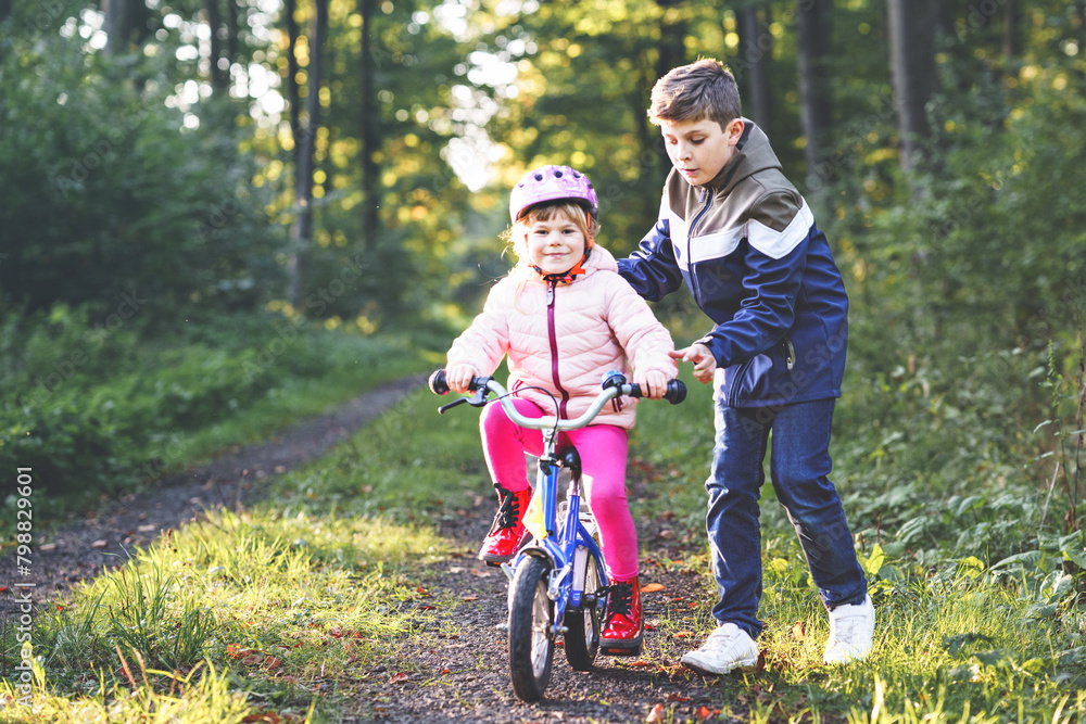 Cute little preschool girl in safety helmet riding bicycle. School kid boy, brother teaching happy healthy sister child cycling and having fun with learning bike. Active siblings family outdoors. Cute little preschool girl in safety helmet riding bicycle. School kid boy, brother teaching happy healthy sister child cycling and having fun with learning bike. Active siblings family outdoors.