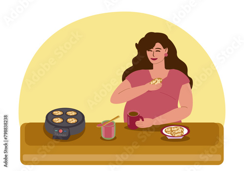 A pregnant young woman has breakfast, drinks tea and eats pancakes with jam. There is an electric pancake maker and a jar of jam on the table. Motherhood, pregnancy, healthy eating. 