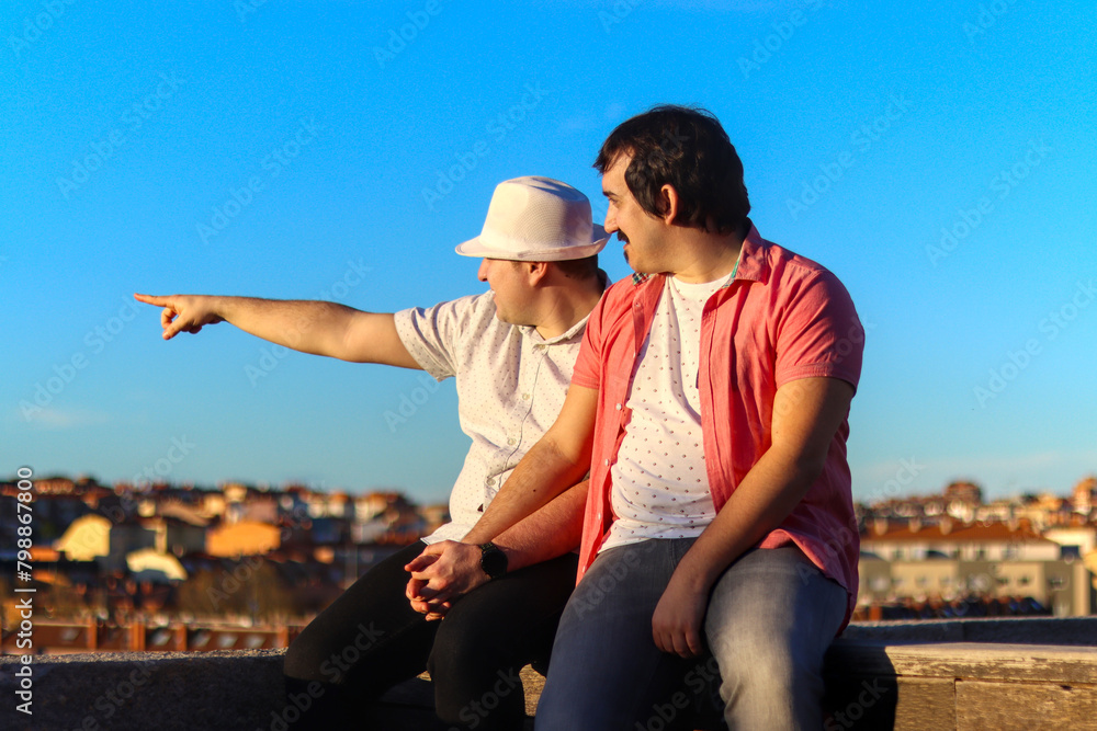 Couple of gay men in a lookout point, pointing to the left horizon with the city in the background. LGBT love concept