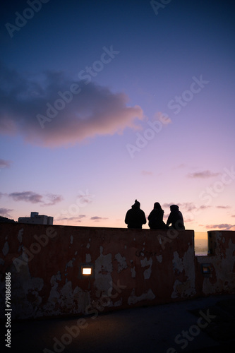 Three person silhouette sitting on a wall in a sunset